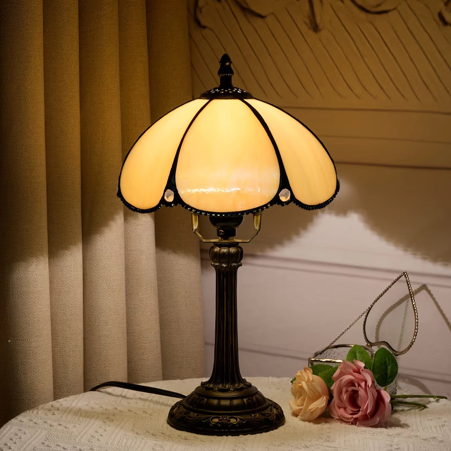 A vintage-style table lamp with a warm yellow stained glass shade and a dark bronze metal base, placed on a white lace-covered table next to pink roses in front of beige curtains.