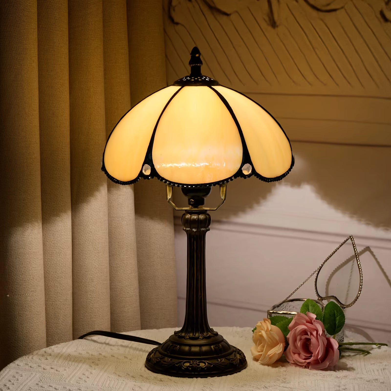 A vintage-style table lamp with a warm yellow stained glass shade and a dark bronze metal base, placed on a white lace-covered table next to pink roses in front of beige curtains.