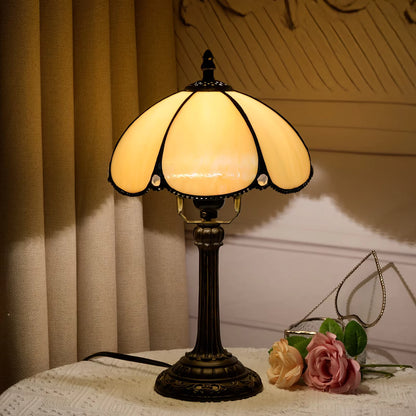 A vintage-style table lamp with a warm yellow stained glass shade and a dark bronze metal base, placed on a white lace-covered table next to pink roses in front of beige curtains.