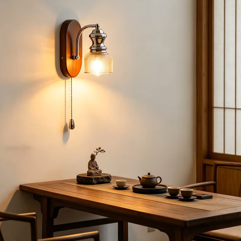 A warm wall-mounted sconce with a glowing glass shade and pull chain, installed above a wooden table in a softly lit interior with shoji-style window panels.