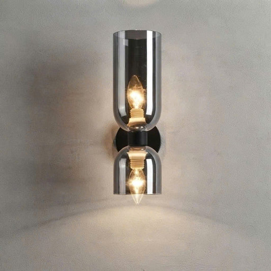 A modern wall-mounted sconce with two mirrored glass shades and glowing warm bulbs, set against a textured light gray wall.