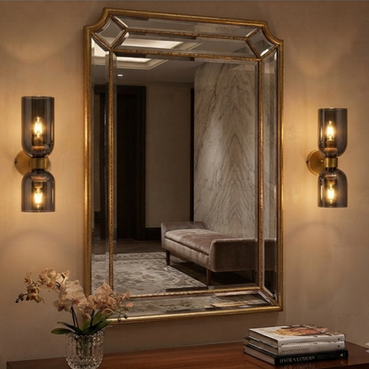 A large rectangular wall mirror with an ornate gold frame hangs on a beige wall, flanked by two matching gold and smoked glass sconces. The mirror reflects a furnished interior with a bench and marble wall, while a wooden console table with books and a vase of flowers sits below.