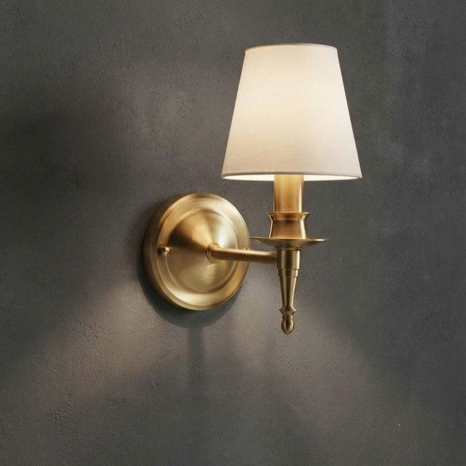 A single wall-mounted sconce with a warm brass/gold metal fixture and a tapered off-white fabric shade, mounted on a dark textured wall.