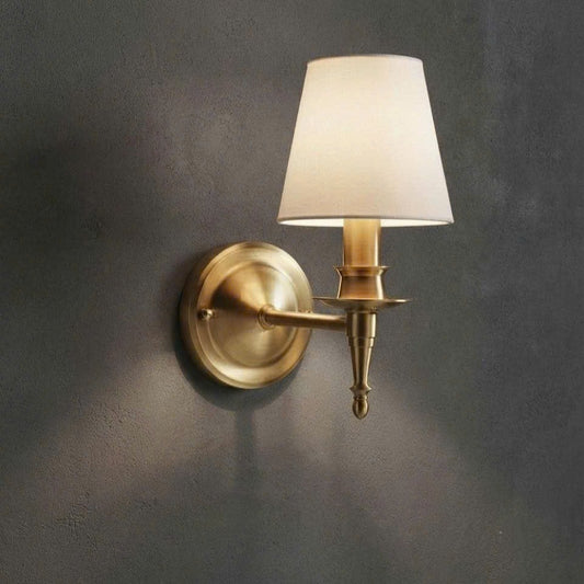 A single wall-mounted sconce with a warm brass/gold metal fixture and a tapered off-white fabric shade, mounted on a dark textured wall.
