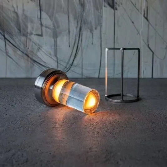 A modern cylindrical wall light with a glowing amber LED inside a clear glass or acrylic tube, mounted on a dark metal base, shown next to a matching rectangular metal frame on a concrete floor against an abstract painted wall.